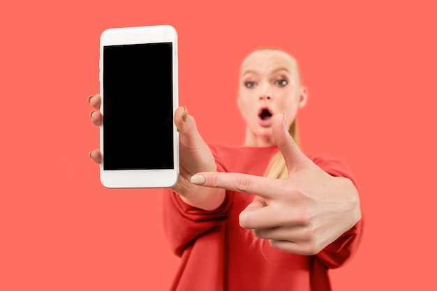 Free photo portrait of a surprised, smiling, happy, astonished girl showing blank screen mobile phone isolated over coral background.