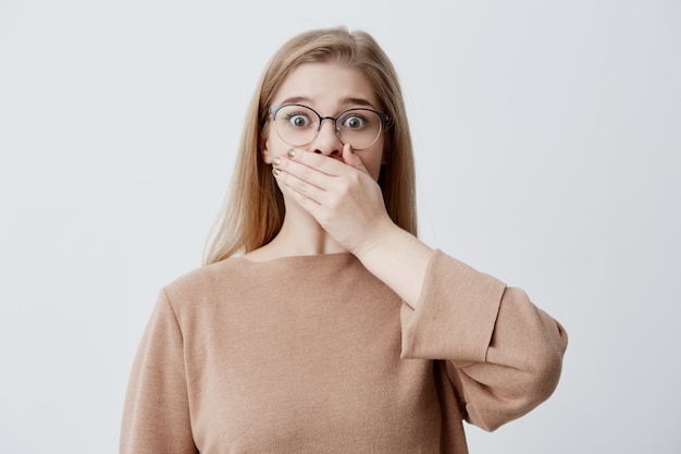 Portrait of surprised worried young woman in spectacles covers mouth with hand, tries to keeps secret and not to spread gossips or confidential information. People and silence concept