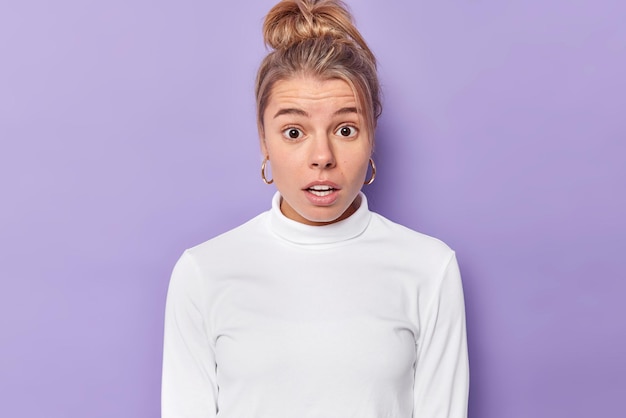 Free photo portrait of surprised young european woman stares with big eyes holds breath cannot believe her eyes wears white poloneck isolated over purple background feels fascinated hears stunning news.