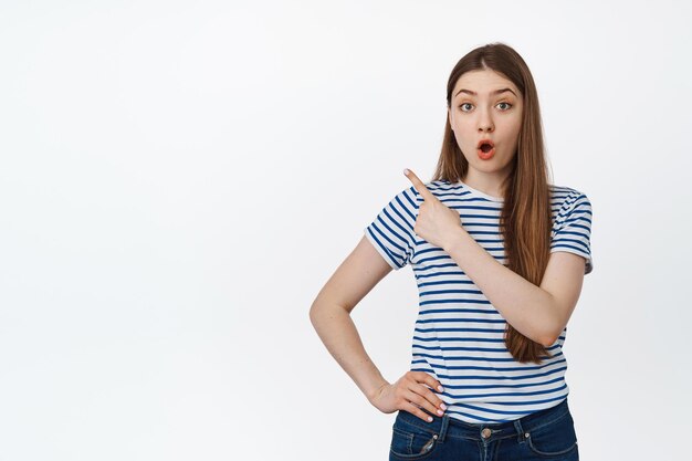 Portrait of surprised young woman pointing at upper left corner, showing exciting announcement, standing over white background