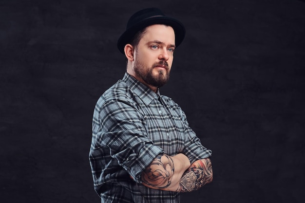 Portrait of a tattoed middle age hipster man with beard and hairstyle dressed in a checkered shirt and hat, pose with crossed arms in a studio. Isolated on a dark textured background.