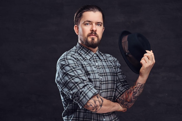 Portrait of a tattoed middle age hipster man with beard and hairstyle dressed in a checkered shirt pose with a hat in hand. Isolated on a dark textured background.