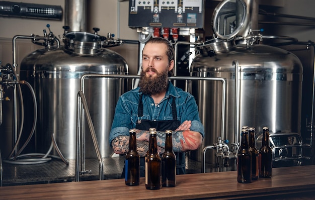 Portrait of tattooed, bearded hipster male manufacturer presenting beer in the microbrewery.