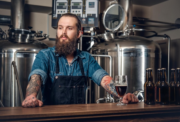 Portrait of tattooed, bearded hipster male manufacturer presenting beer microbrewery.