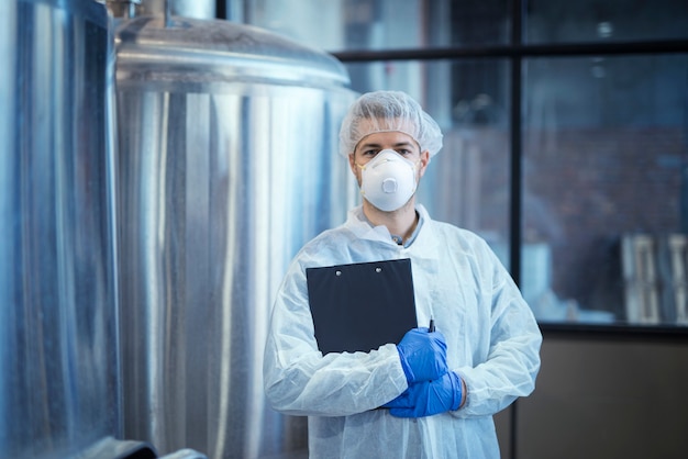 Portrait of technologist in white uniform with hairnet and protective mask and gloves standing in pharmaceutical or food factory with arms crossed