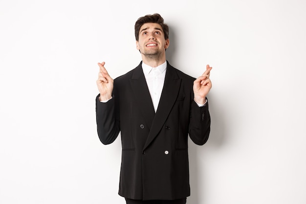 Free photo portrait of tensed and worried handsome businessman, crossing fingers and looking up, begging god, making a wish, standing against white background in black suit.