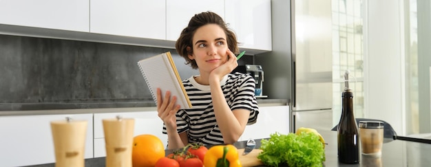 Free photo portrait of thinking woman with notebook cooking writing down recipe ingredients deciding on a meal