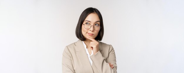 Portrait of thoughtful asian businesswoman in glasses making assumption thinking standing in beige suit against white background