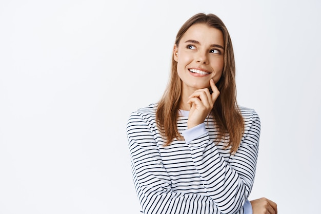 Free photo portrait of thoughtful blond woman having an idea, looking pensive at upper left corner, imaging something good, standing against white wall