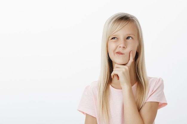 Portrait of thoughtful creative young girl with long blond hair, looking up and holding hand on chin while thinking or calculating in mind