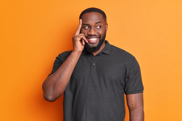 Portrait of thoughtful handsome bearded man keeps finger on temple smiles pleasantly thinks about decision looks away dressed in casual black t shirt isolated over orange wall