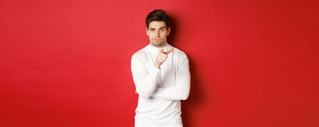 Portrait of thoughtful handsome man in white sweater, pointing at camera and making choice, standing against red background.