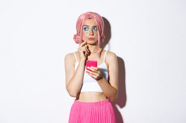 Portrait of thoughtful young woman in pink wig, with bright makeup, looking aside while thinking, holding mobile phone, standing over white background