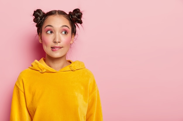 Portrait of thoughtful young woman with two hair buns, bites lips, being deep in thoughts, tries to find solution in mind, dressed in yellow jumper