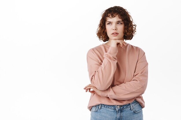 Portrait of thoughtul curly girl, student making assumption, look serious and thinking, staring at upper left corner focused, pondering decision, standing on white