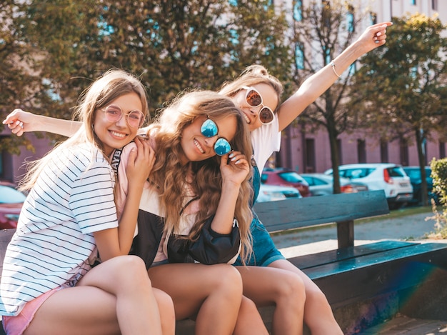 Free Photo portrait of three young beautiful smiling hipster girls in trendy summer clothes.sexy carefree women sitting on the bench in the street.positive models having fun in sunglasses.raising hands