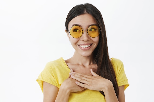 Portrait of touched and pleased blushing european female in trendy yellow sunglasses holding palms in delight