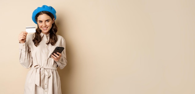 Free Photo portrait of trendy woman in coat holding smartphone showing credit card shopping online order someth