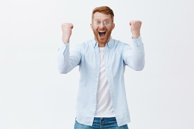 Free photo portrait of triumphing happy caucasian male with ginger hair and bristle in glasses yelling from success and positive emotions raising fists to celebrate victory