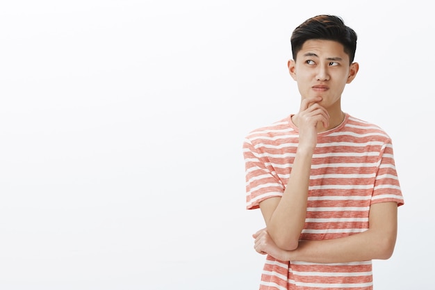 Portrait of troubled young asian guy trying think up plan or idea, standing in thoughtful pose with hand on chin, looking questioned and hesitant at upper left corner