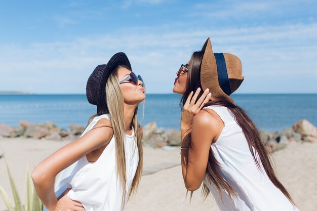 Portrait of two attractive brunette and blond girls with long hair are standing on the beach near sea. They shows a kiss.