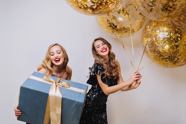 Portrait two joyful excited gorgeous women with long curly hair celebrating birthday party on white space. Big present, balloons with golden tinsels,