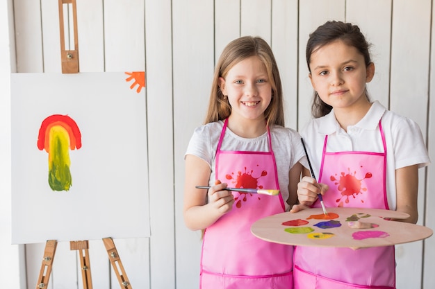 Free photo portrait of a two smiling girls in pink apron looking at camera while painting on the easel