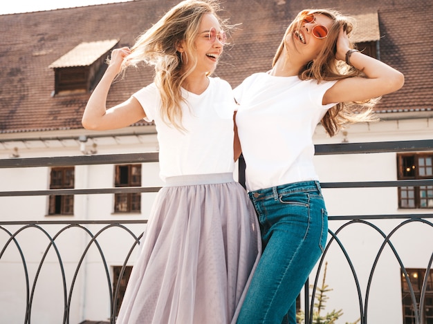 Portrait of two young beautiful blond smiling hipster girls in trendy summer white t-shirt clothes.   . Positive models having fun in sunglasses.Hugging