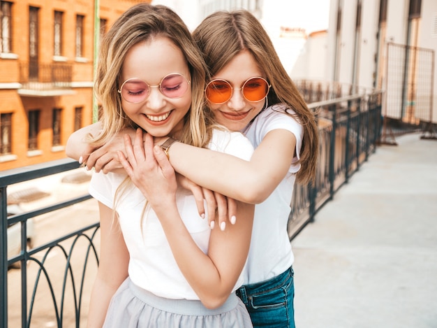 Free photo portrait of two young beautiful blond smiling hipster girls in trendy summer white t-shirt clothes.   . positive models having fun in sunglasses.hugging