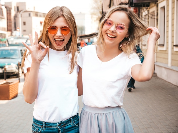 Portrait of two young beautiful blond smiling hipster girls in trendy summer white t-shirt clothes.   . Positive models having fun in sunglasses.Hugging