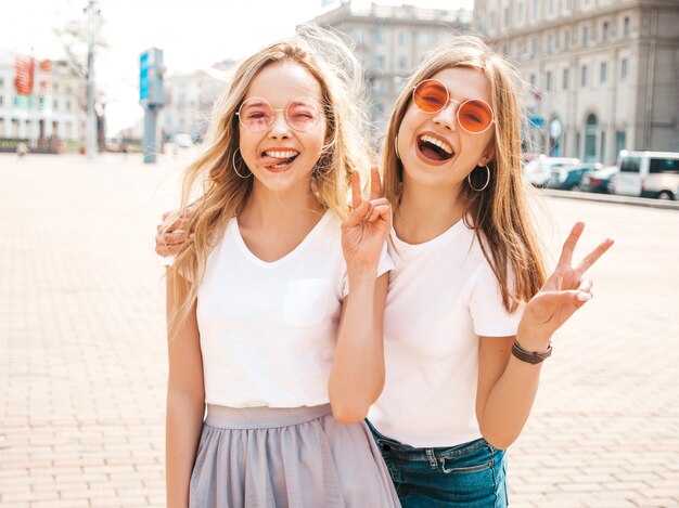 Portrait of two young beautiful blond smiling hipster girls in trendy summer white t-shirt clothes. Sexy carefree women posing on street  . Positive models showing peace sign and tongue