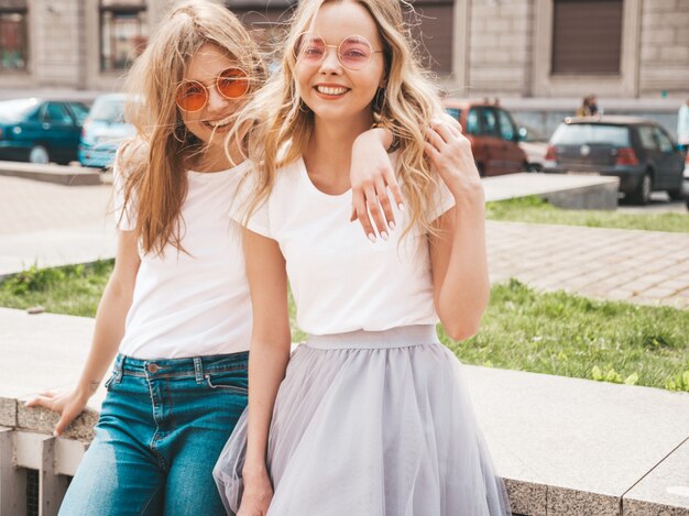 Portrait of two young beautiful blond smiling hipster girls in trendy summer white t-shirt clothes.  