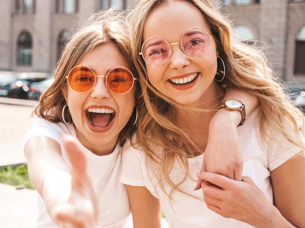 Portrait of two young beautiful blond smiling hipster girls in trendy summer white t-shirt clothes.  