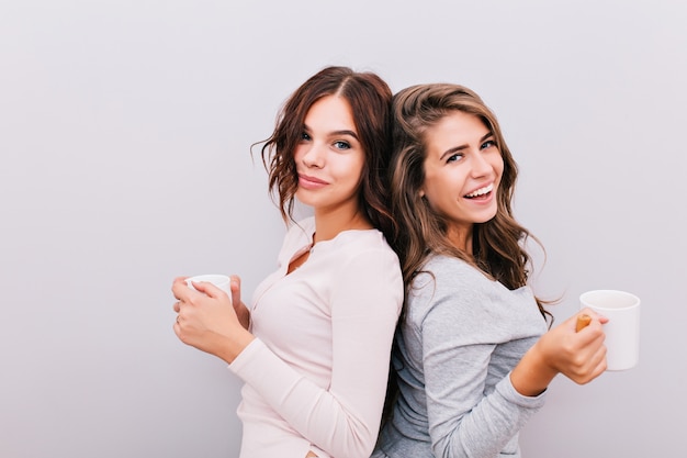 Portrait two young girls in pajamas with cups on gray wall . They stand back to back and smiling .