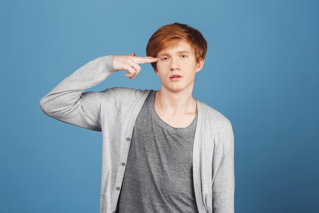 Free photo portrait of unhappy attractive ginger male student in casual gray outfit holding hand in gun gesture near hand, being tired and stressed with lot of tasks on study, having bad mood.