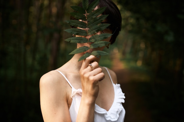 Portrait of unrecognizable mysterious young female wearing white strap dress posing in forest alone covering face with large fern leaf