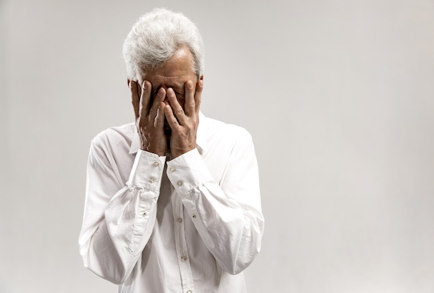 Portrait of upset old man covering face while crying. Isolated on grey wall