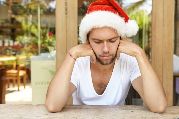 Portrait of upset young male dressed in white t-shirt and red Santa Claus hat feeling sick after boozing with friends at New Year's party