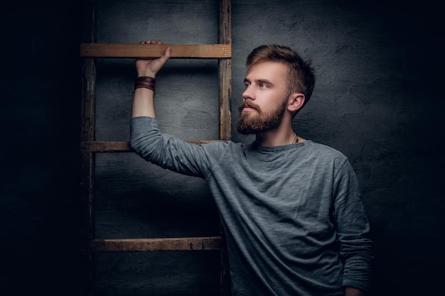 Free photo portrait of an urban bearded male posing near old vintage ladder on grey background.