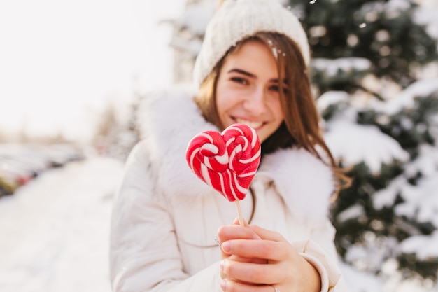 Portrait winter young woman holding pink heart lollypop, chilling on street full with snow in sunny morning. White knitted hat, smiling.