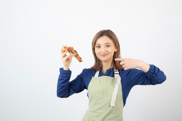 Portrait of woman in apron pointing at slice of pizza on white 