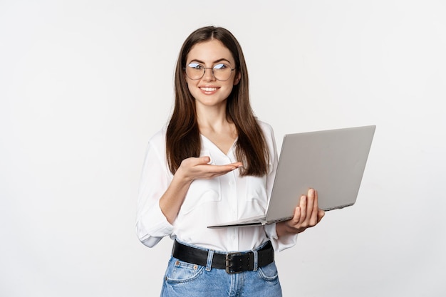 Portrait of woman in glasses holding laptop pointing at screen showing her work on computer standing...