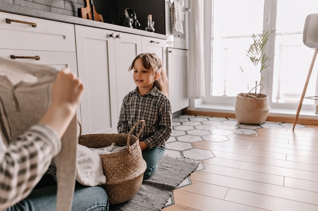 Free photo portrait of woman in plaid shirt sitting on floor of kitchen and watching her mother take out clothes from basket.