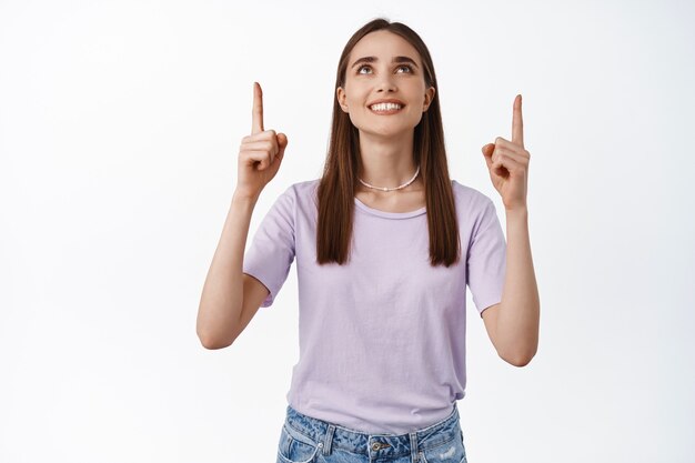 Portrait of woman pointing and looking up with satisfied white smile making her choice, standing on white in t-shirt