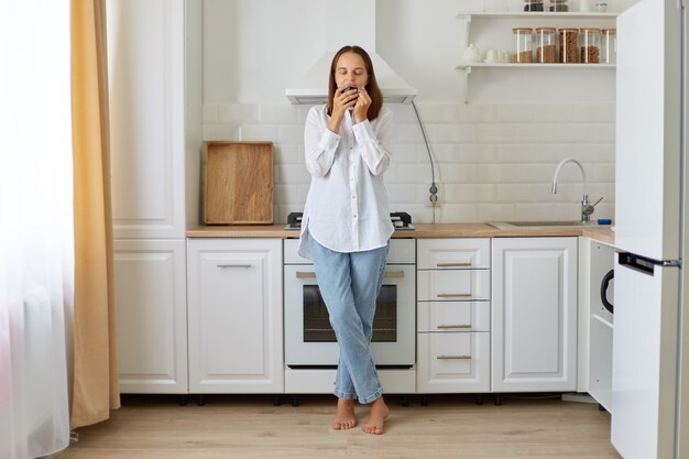 Portrait of a woman wearing white shirt and jeans standing smelling a cup of coffee in her kitchen, smelling aroma beverage in morning while posing in kitchen.