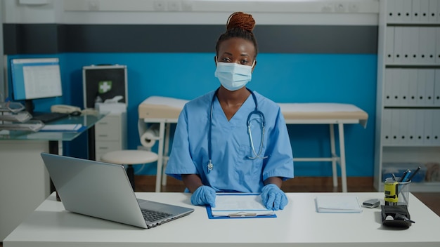 Portrait of woman working as medical assistant wearing face mask and uniform while sitting at healthcare clinic. Nurse at desk with laptop and files looking at camera in hospital room