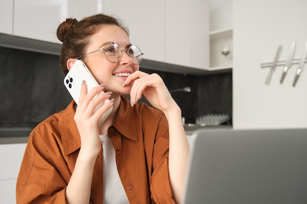 Free Photo portrait of woman working from home with laptop making a phone call using smartphone looking