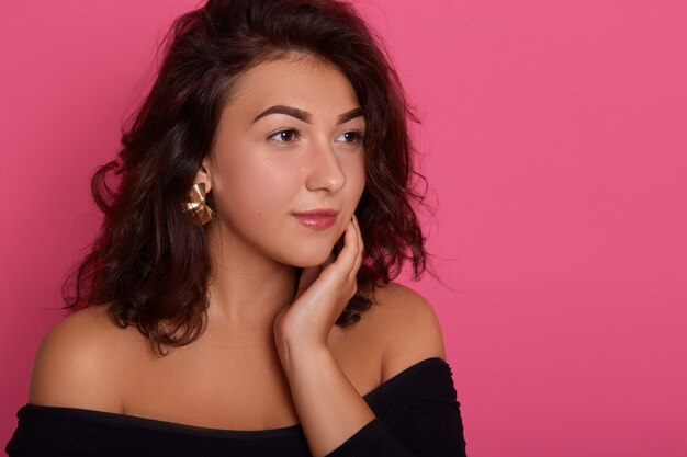 Portrait of wonderful young brunette woman looking dreamily aside, keeping hand on her neck, posing with bare shoulders, dark haired girl with earrings.