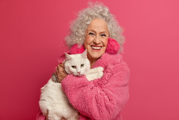 Free Photo portrait of wrinkled grandmother holds white cat on hands, stays at home during pandemic outbreak, wears fluffy earrings, soft robe, going to feed pet, isolated on pink wall. woman on pension