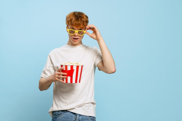 Portrait of yong emotive boy in 3D glasses with popcorn basket posing isolated over blue studio backgound Movie time